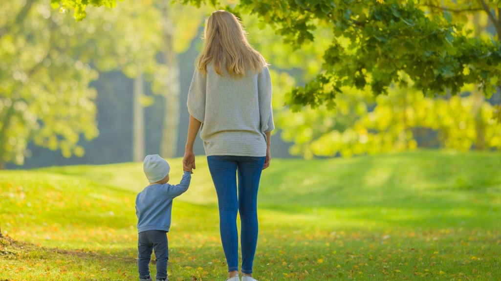 Young adult mother and baby boy standing on grass at city park and looking far away. Spending time together in beautiful warm sunny autumn day. Back view.