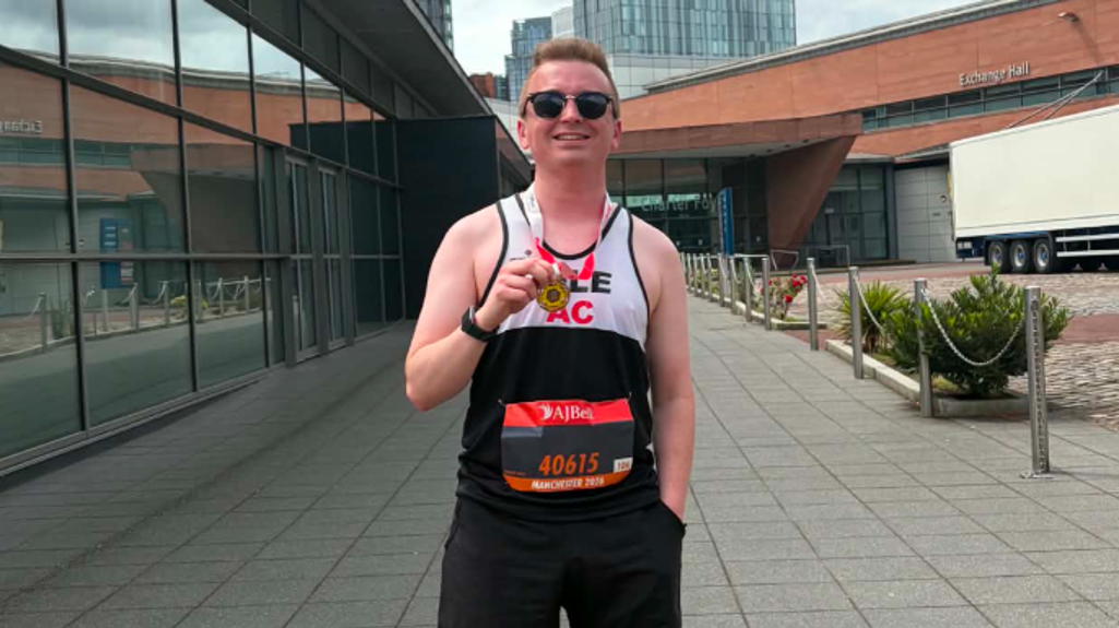 Liam stands on a paved walkway between modern buildings, wearing running gear, holding the medal up toward the camera under a cloudy sky.