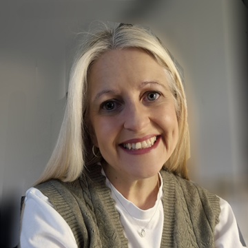 A person with long, light-coloured hair is seated indoors, wearing a white long‑sleeved top layered under an olive‑green knitted vest with a cable pattern. The individual has arms folded, with a silver bracelet visible on one wrist. Soft natural light illuminates the scene against a plain background.