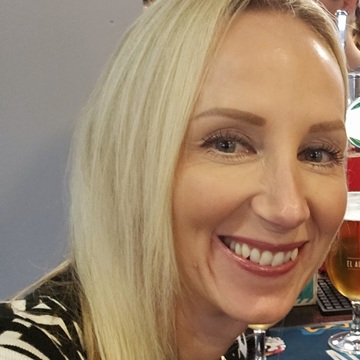 A person with long, straight blonde hair is seated indoors at what appears to be a pub. They are wearing a black-and-white patterned top with gold buttons on the shoulders. 