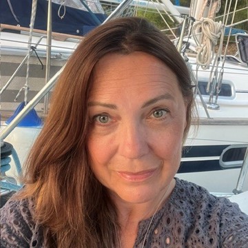 Clare Harrigan smiling at the camera outdoors beside a marina. She is wearing a grey patterned top and standing in front of several sailboats. The photo is taken in natural evening light.