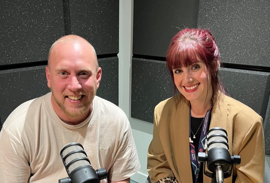 Joe, a man with a trimmed beard, wearing a light beige T-shirt, and Sian, a woman with dark red hair tied back, wearing a tan blazer over a black top with leopard-print detail on the sleeves, sit side by side in a recording studio, smiling at the camera with microphones in front of them.