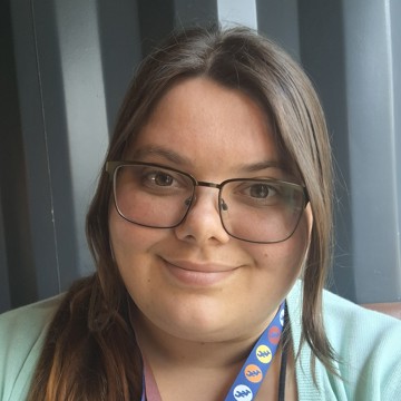 A person with long brown hair is seated indoors against a dark, vertically‑panelled wall. The person is wearing a light‑coloured top and a lanyard with a patterned design featuring small circular icons in blue, orange, and yellow. 
