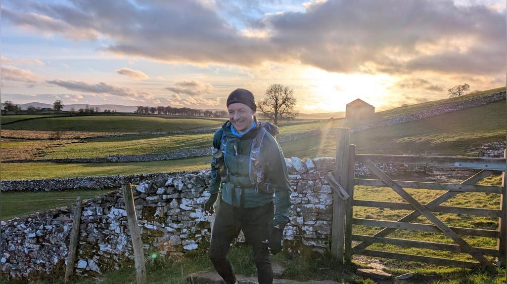 Richard walking through a countryside setting at sunset, stepping over a stone stile beside a wooden gate. Richard is wearing outdoor walking gear and a backpack, with rolling green fields, stone walls, and a barn visible in the background under a partly cloudy sky.