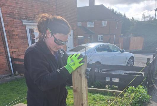 Person wearing safety glasses and gloves installing a wooden fence post into the ground beside a pavement, with string lines marking the fence line.