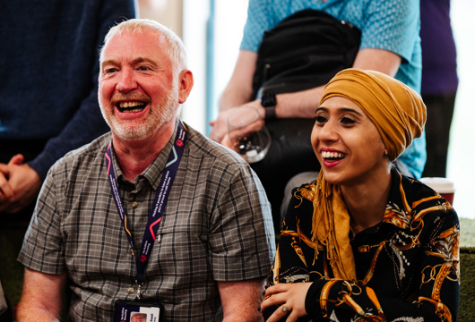 Two people sit side by side in a group setting, appearing to listen attentively. One person wears a checked shirt and a lanyard, while the other wears a patterned outfit with a mustard-yellow head scarf. Other individuals stand or sit in the background, contributing to a casual, event-like atmosphere.
