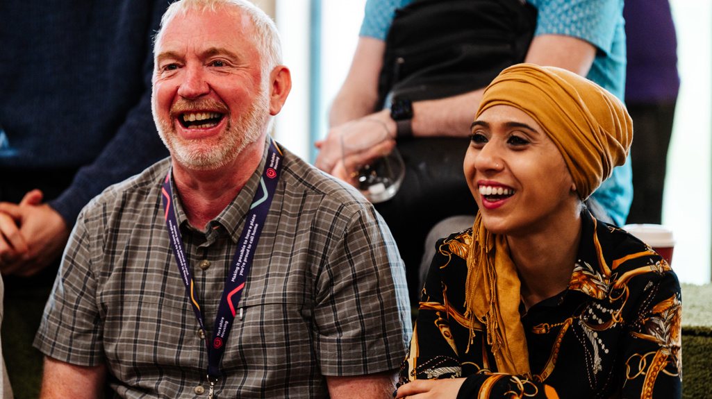 Two people sit side by side in a group setting, appearing to listen attentively. One person wears a checked shirt and a lanyard, while the other wears a patterned outfit with a mustard-yellow head scarf. Other individuals stand or sit in the background, contributing to a casual, event-like atmosphere.