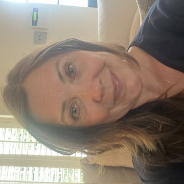 Clare Harrigan smiling at the camera while seated indoors. She is wearing a navy top and is photographed in a bright living room with a cream sofa and window shutters visible in the background.