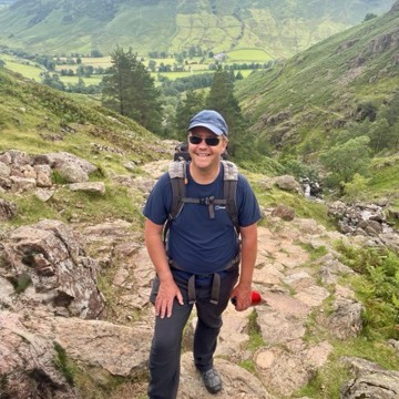 Man smiling at the camera wearing a baseball cap and sunglasses at the top of a big hill after a walk