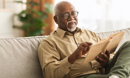 An older gentleman sat on a sofa reading a book