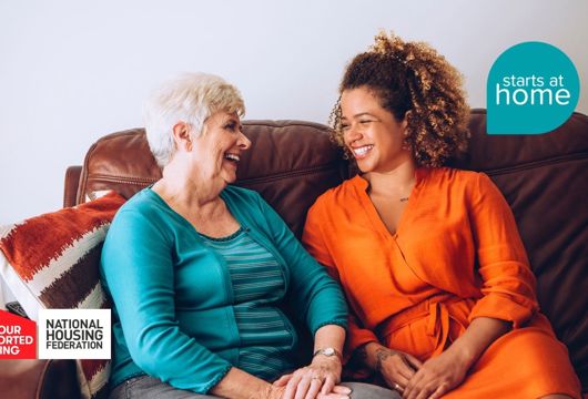 Two women sitting on a sofa, smiling warmly at each other and holding hands. The image includes logos for 'Save Our Supported Housing,' 'National Housing Federation,' and 'Starts at Home'."