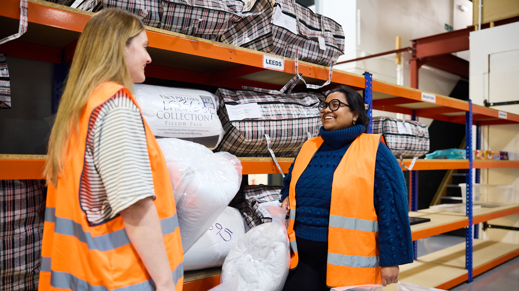 Two volunteers in the Zarach warehouse holding pillows in front of shelves of pillows