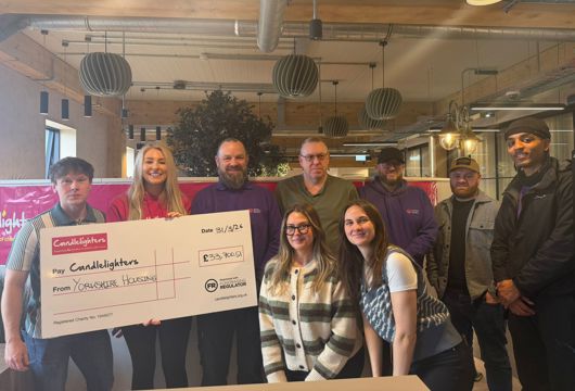 A group of nine people standing indoors in a modern office space, holding a large presentation cheque. The cheque reads “Candlelighters” and “From Yorkshire Housing” with an amount of £33,700.51 and a date of 31/3/26. Hanging lights, exposed ceiling beams, and plants are visible in the background.